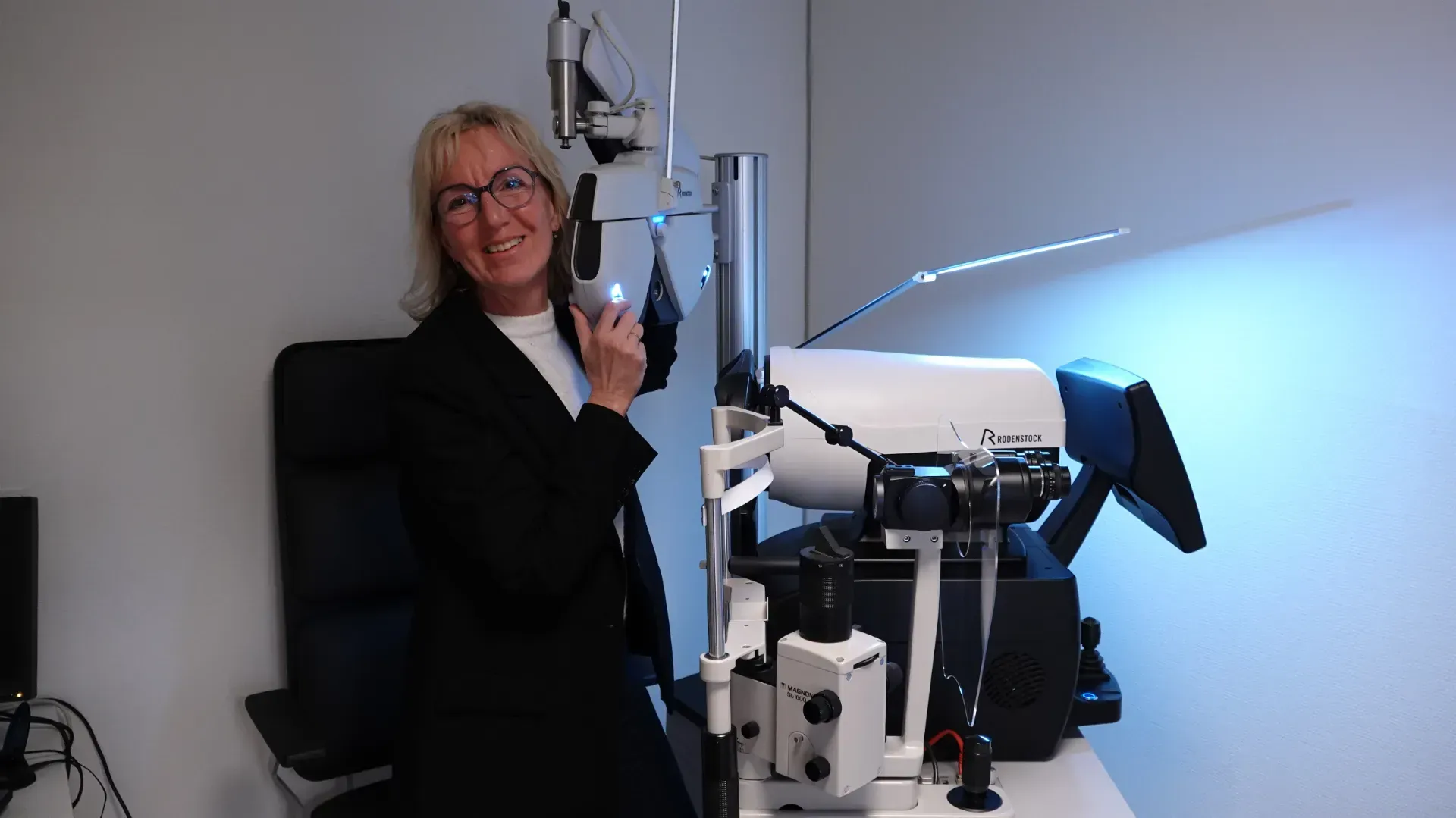 Kvinde med briller smiler og holder en enhed ved siden af ​​en synsundersøgelsesmaskine på en klinik.