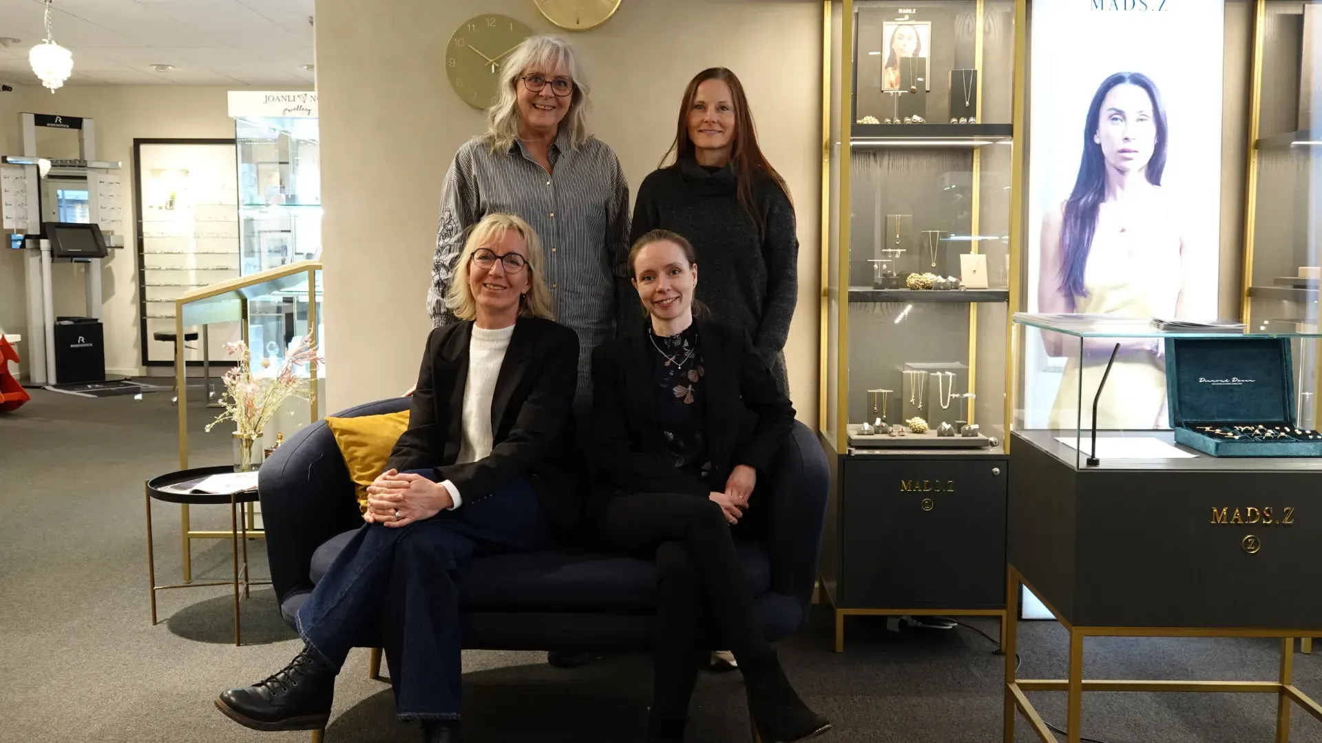 Fire personer poserer i en smykkeforretning. To siddende, to stående, alle smilende.