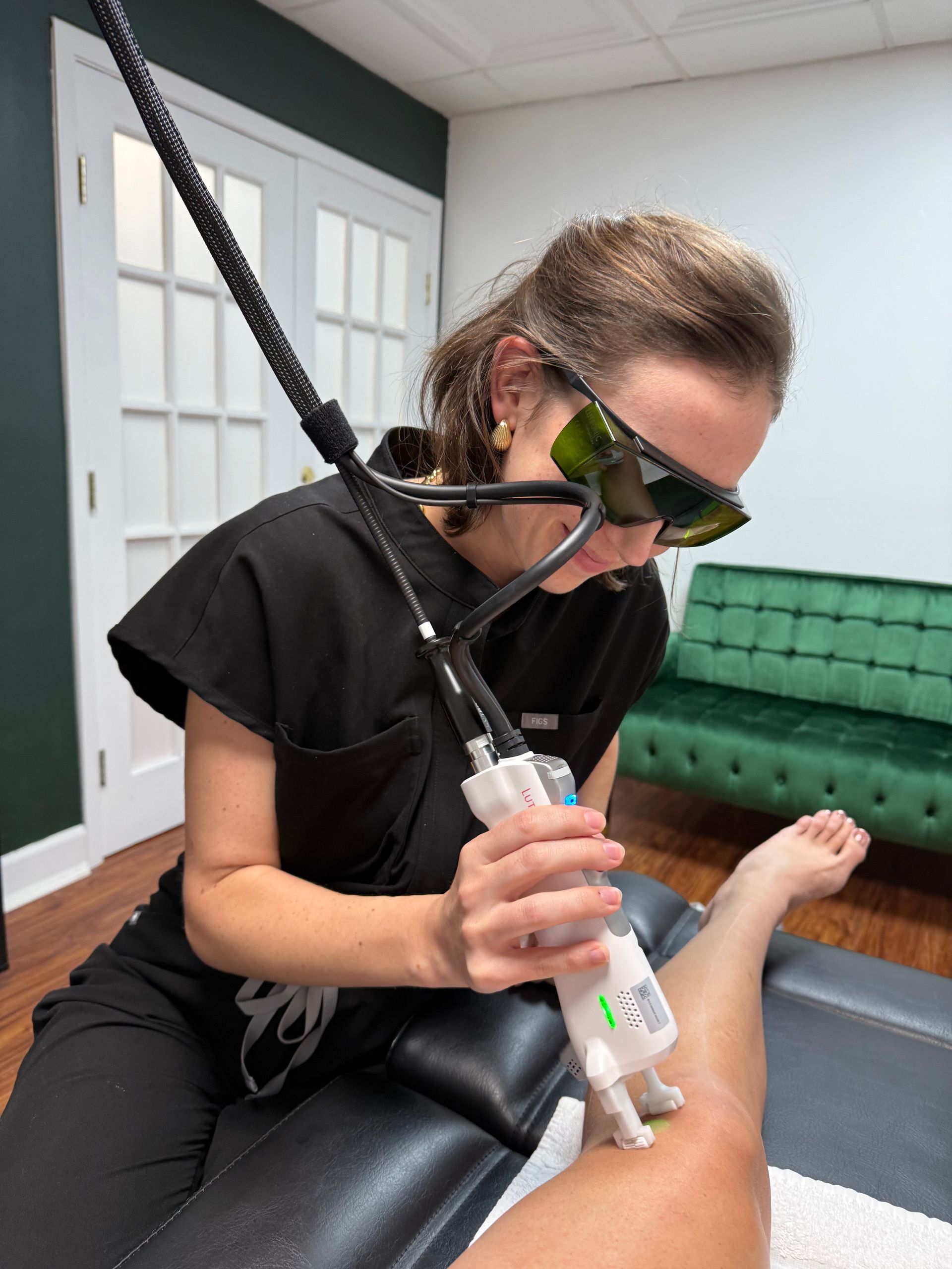 Woman using laser device on a patient's leg in a medical office, wearing safety glasses, black scrubs.