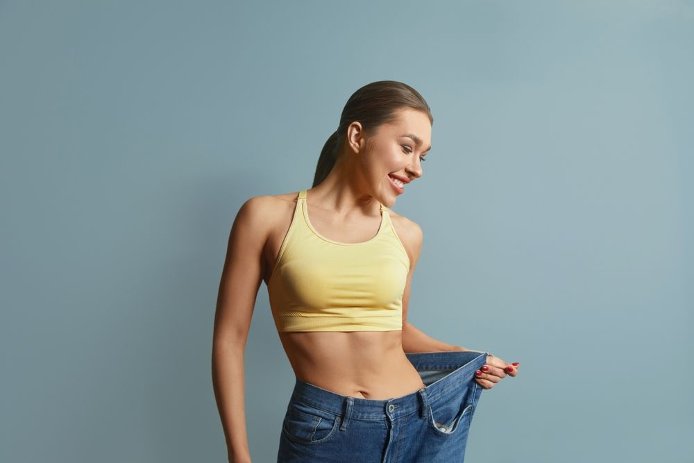 Woman in a yellow sports bra and loose jeans, smiling, holding out the waistband, showing weight loss.