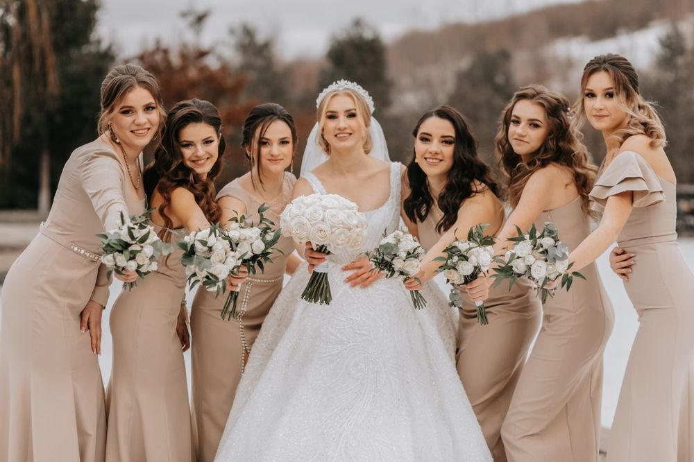 Bride with six bridesmaids in beige dresses holding white bouquets outdoors in a snowy landscape