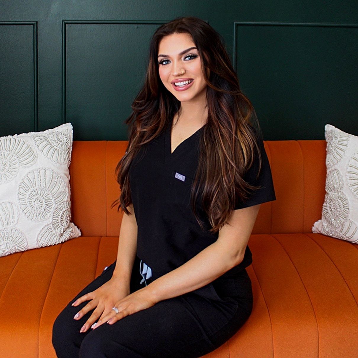 Woman in black scrubs sitting on an orange couch, smiling in a dark green room.