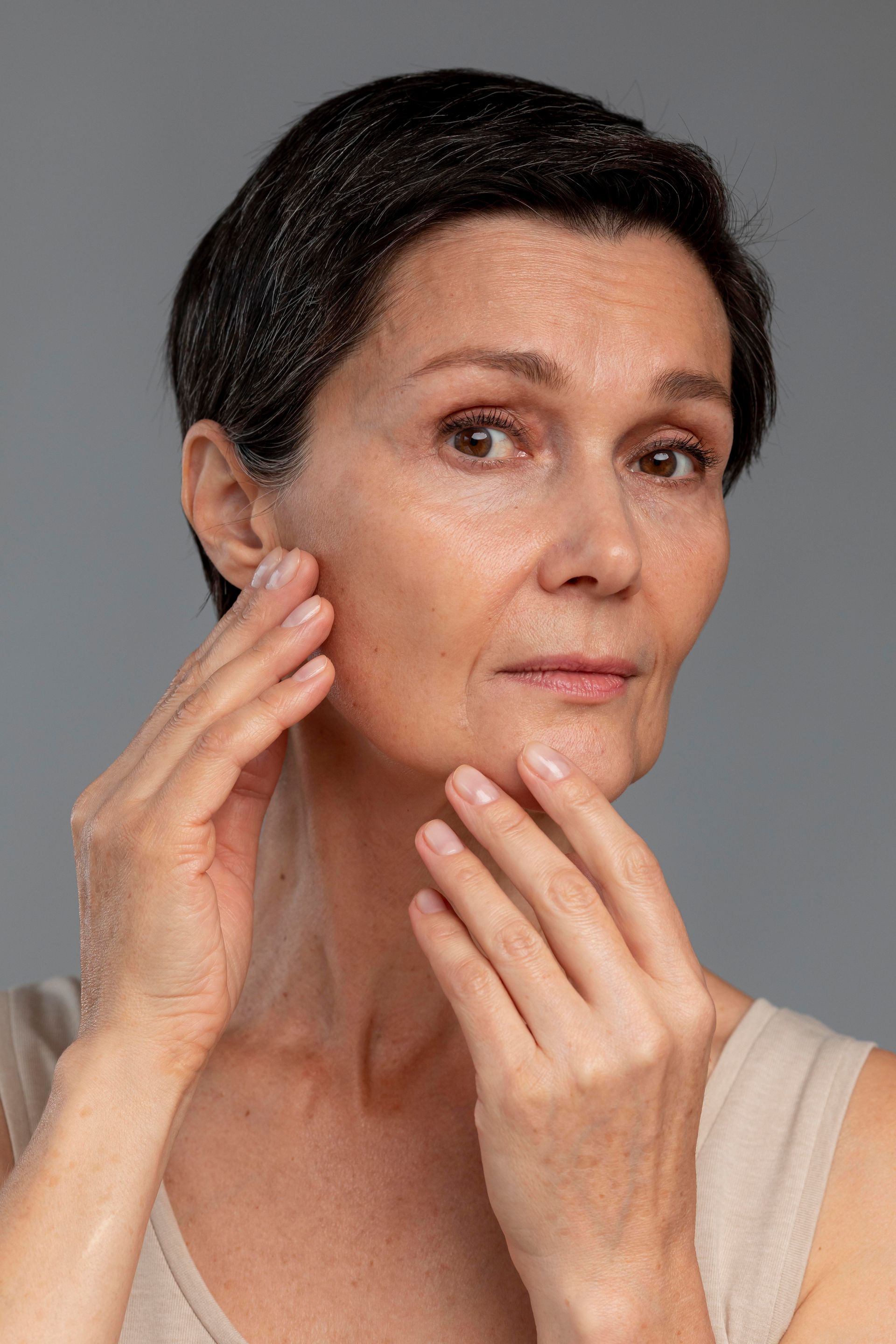 Woman touching face, looking towards the camera, against a gray background.