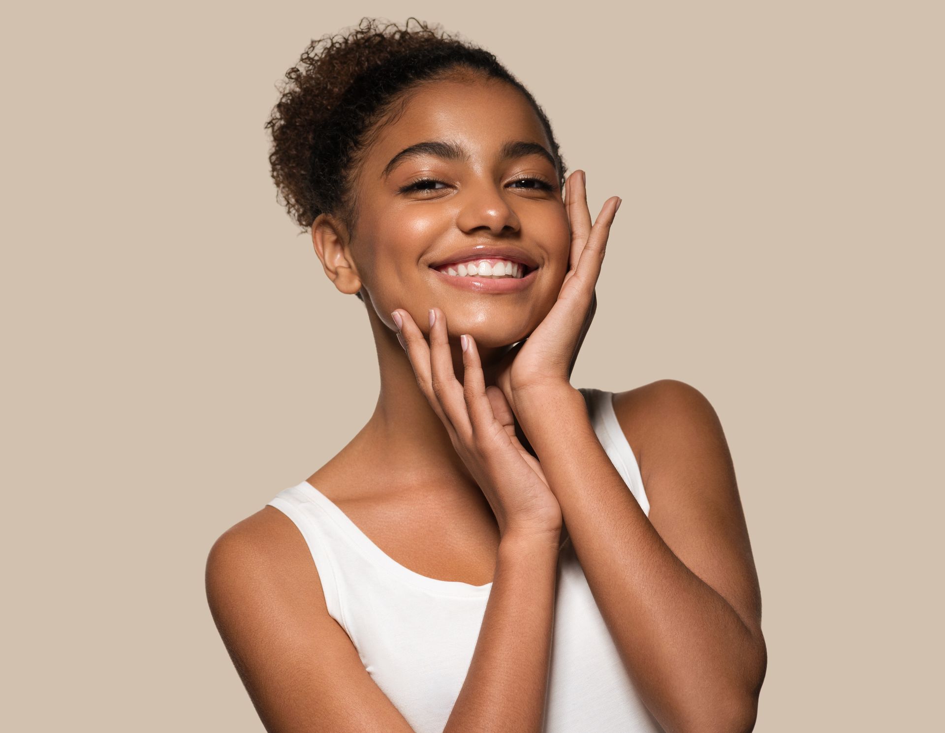 A woman in a white tank top is smiling and touching her face.