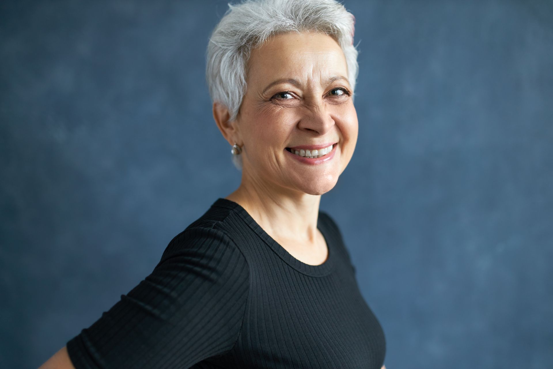 Woman with short gray hair smiles, wearing a black top, against a blue background.