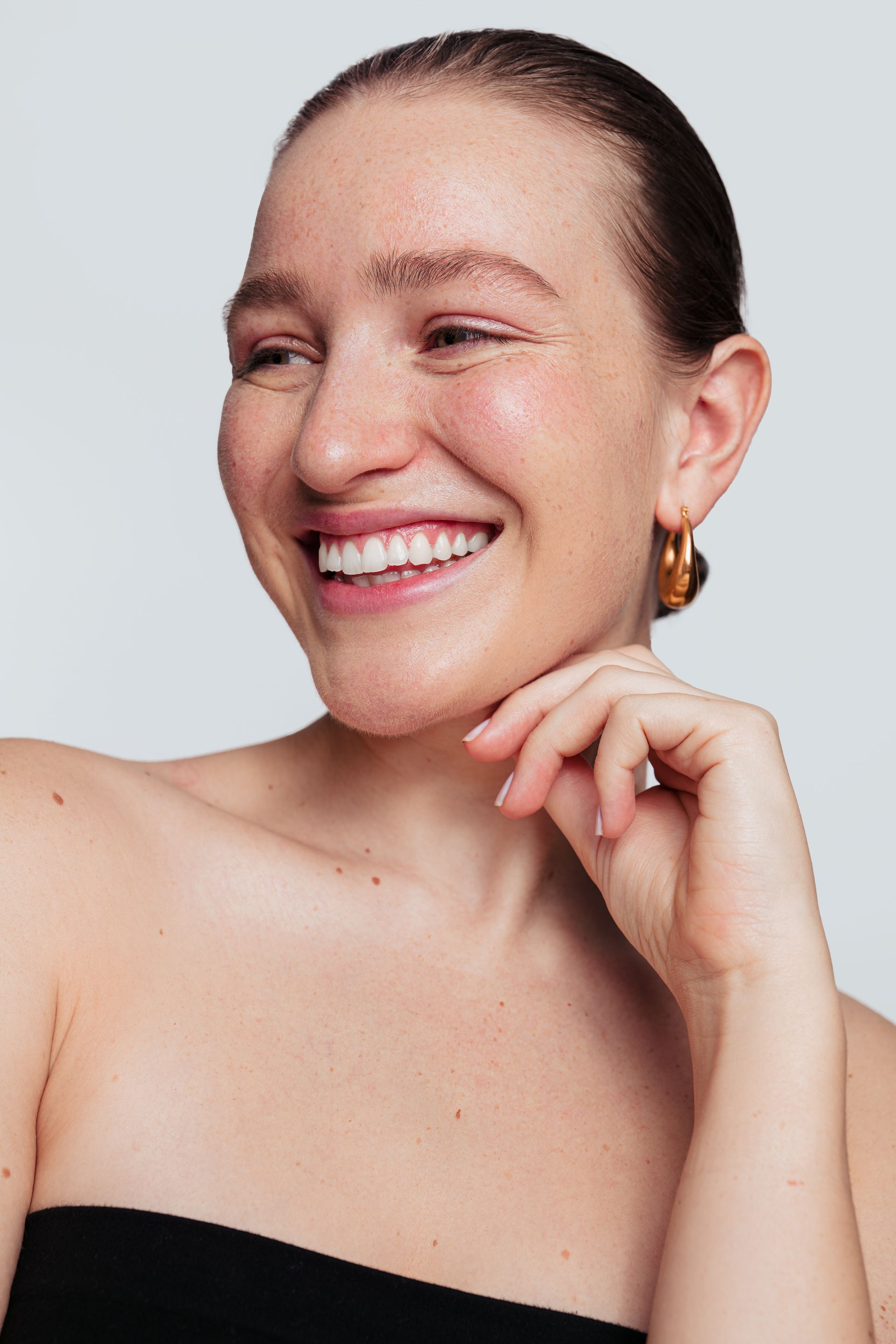 A woman wearing hoop earrings is smiling and touching her face.