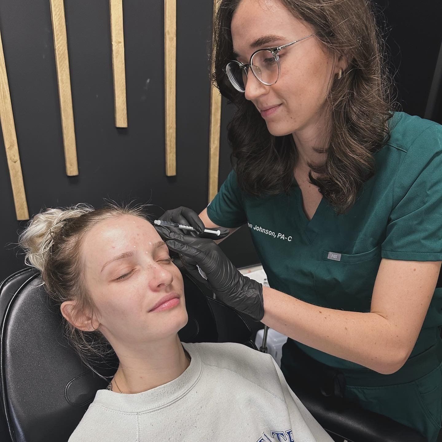 A woman in a green scrub is applying makeup to another woman 's face.