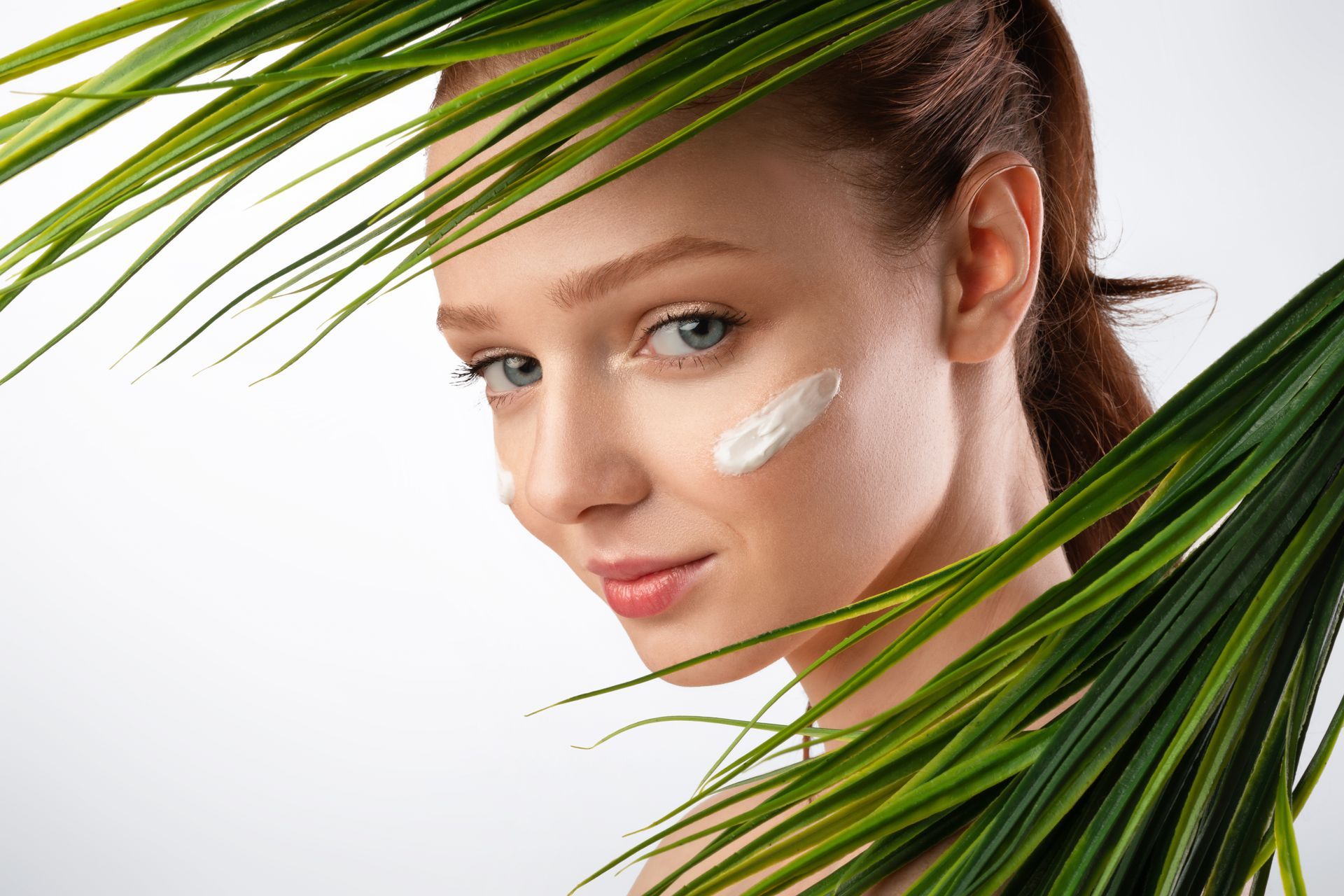 A woman is applying cream to her face behind a palm tree leaf.