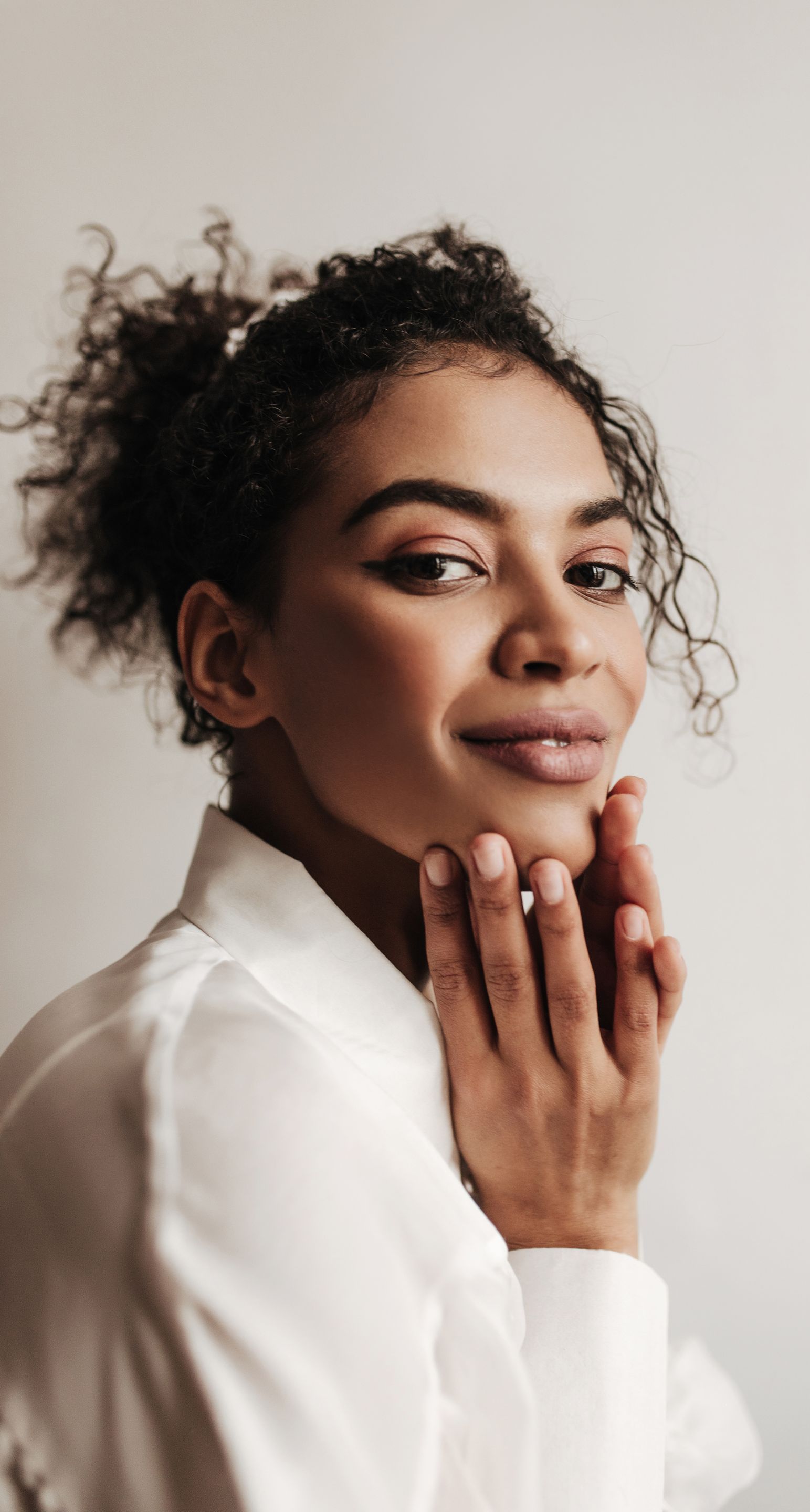 A woman in a white shirt is touching her face and smiling.