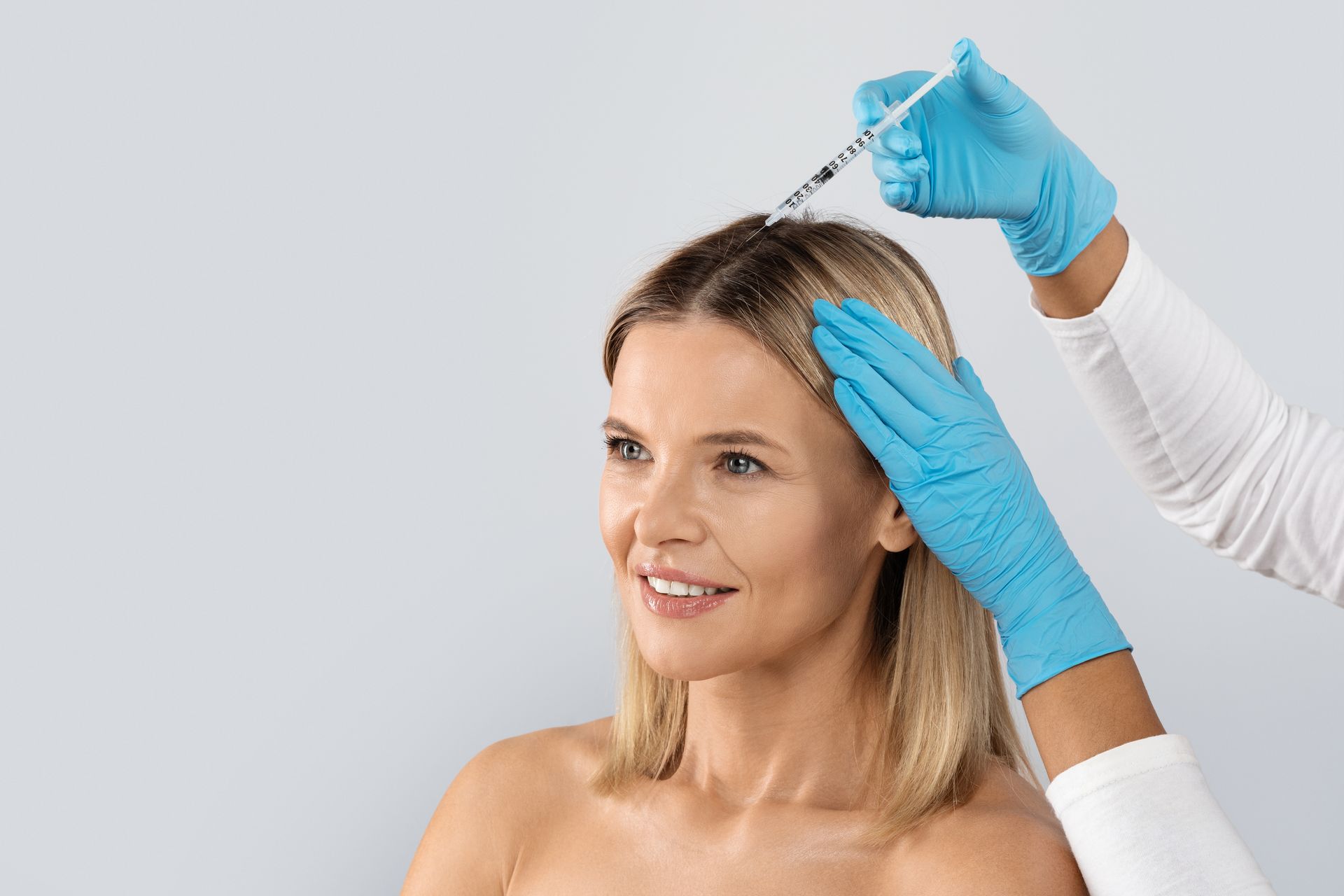 Woman receiving scalp injection, with gloved hands holding syringe, on a light grey background.