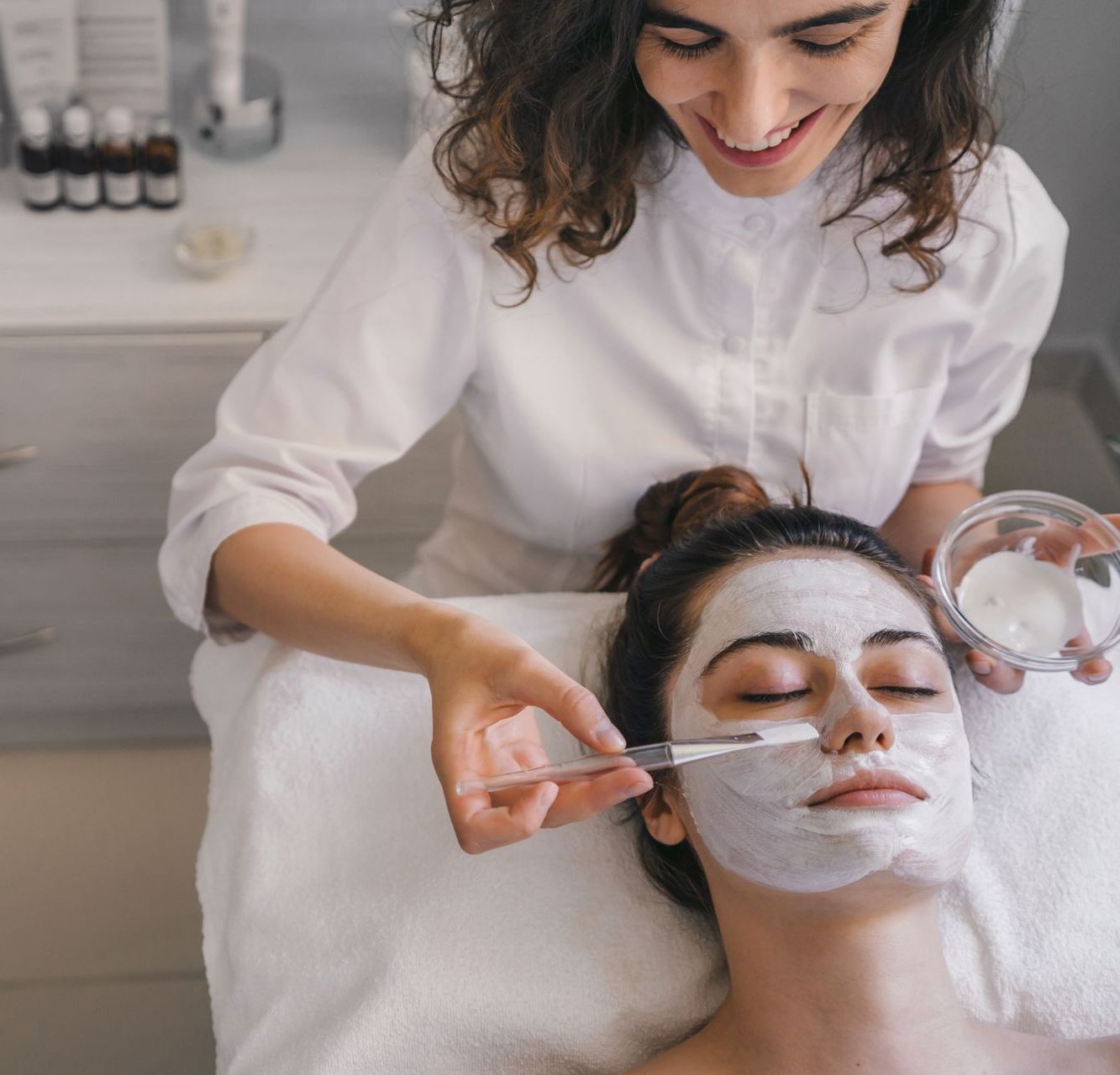 A woman is applying a white mask to another woman 's face.
