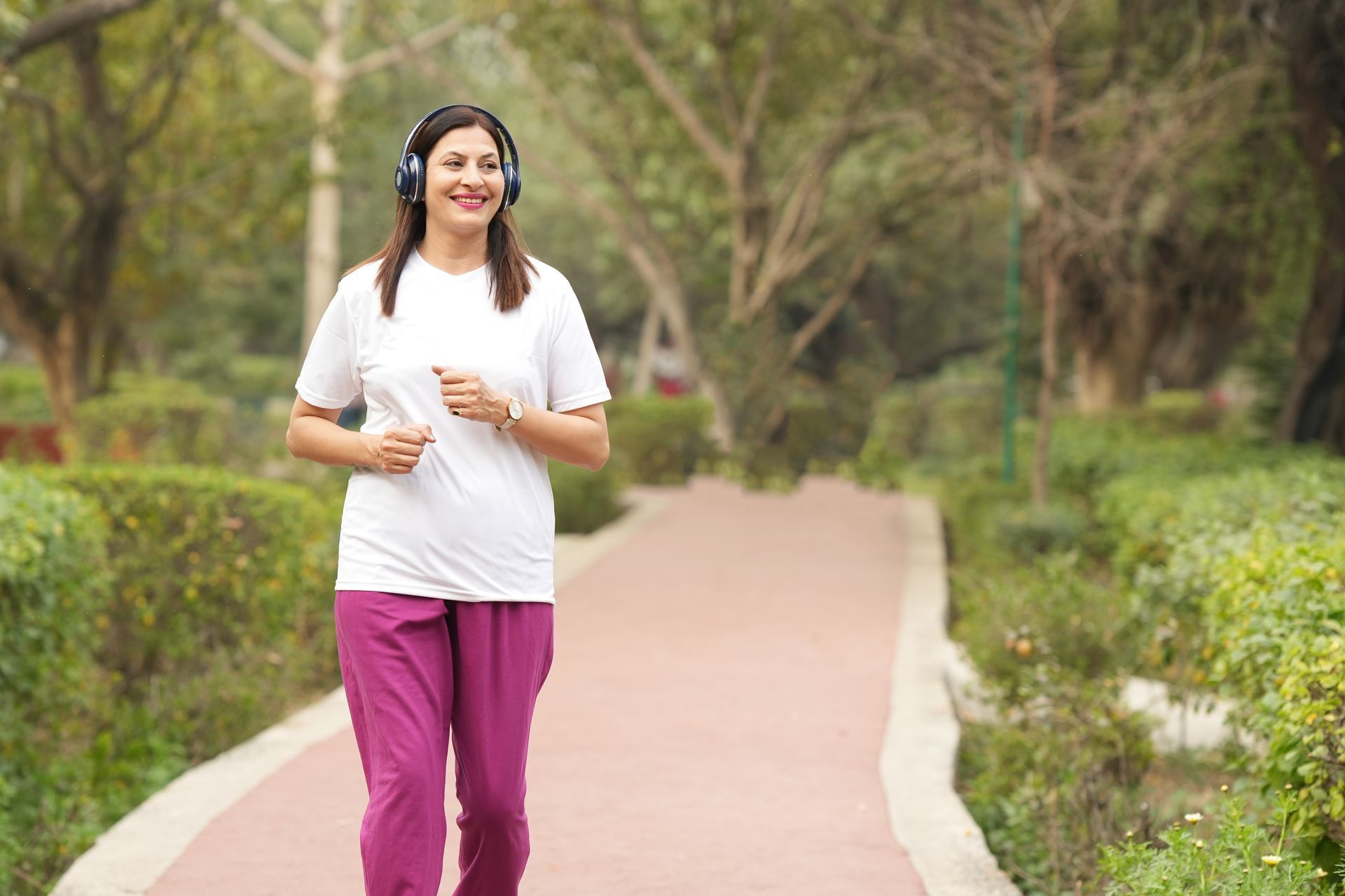 Woman wearing headphones jogs on a path in a park; she wears a white shirt and pink pants.