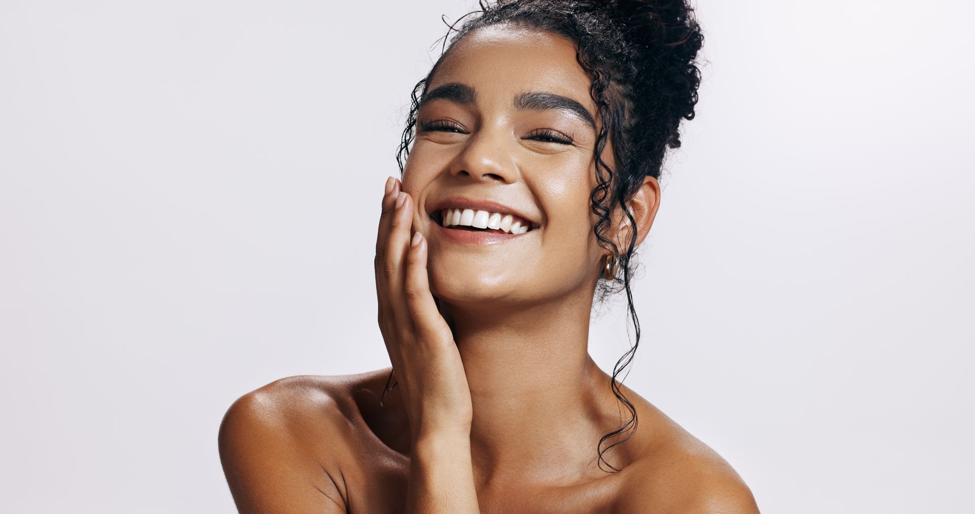 A woman with curly hair is smiling and touching her face.