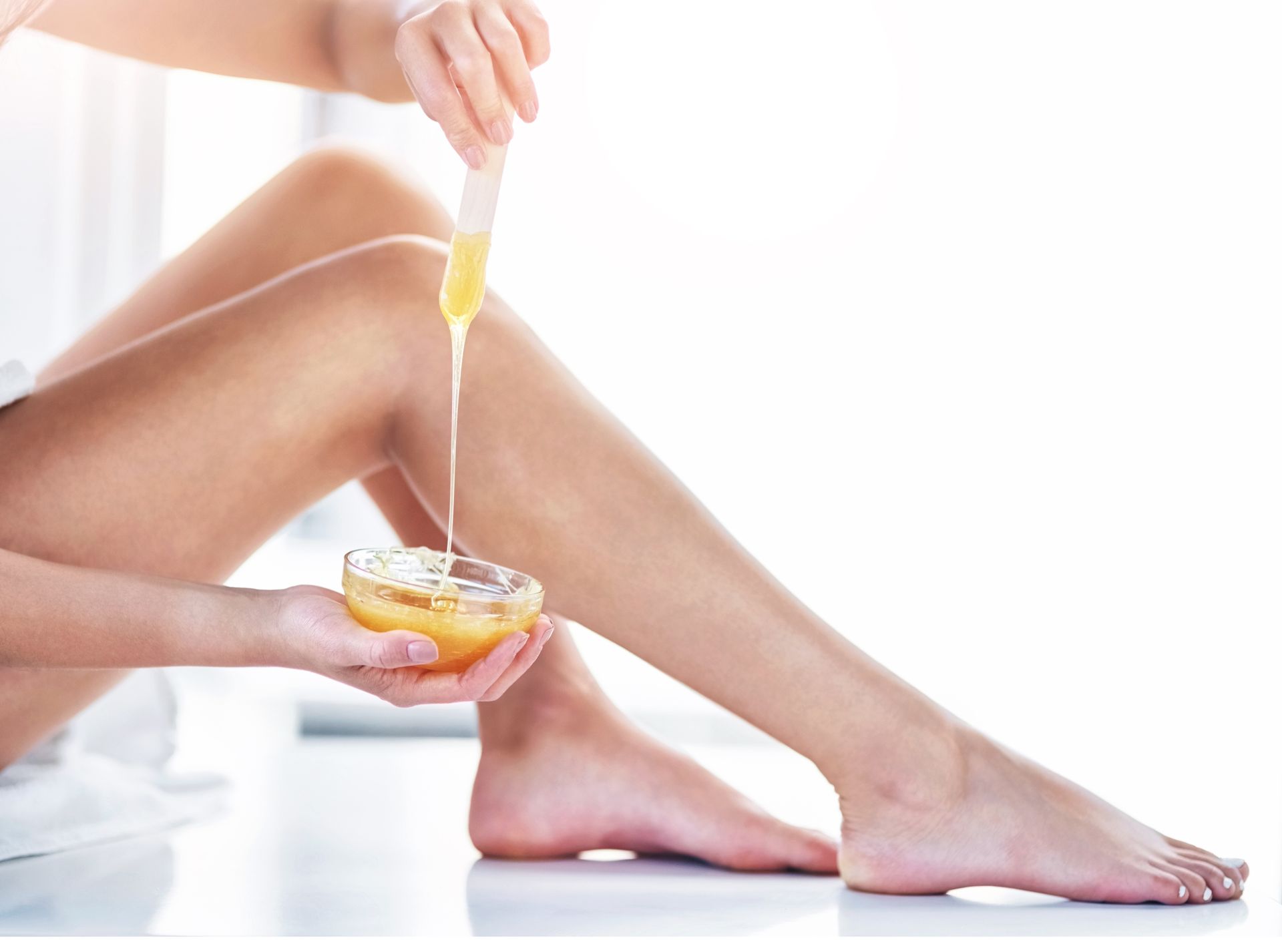 Woman waxing her leg, holding a bowl of wax and applicator stick, in a bright room.