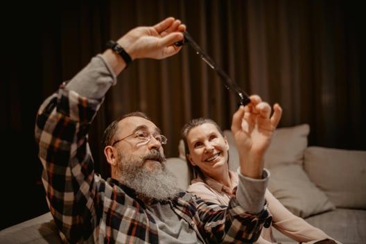 Man and woman looking at a black object. The man holds it up, smiling. Indoors, on a couch.
