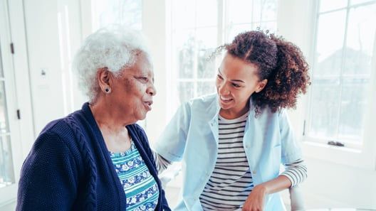 Woman in blue shirt comforts older woman with white hair indoors.