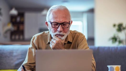 Older adult wearing glasses, using a laptop, resting chin on hand, at home.