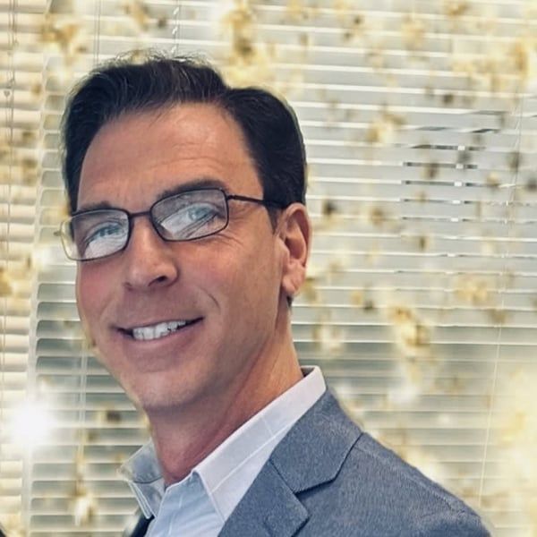 Man wearing glasses and a blazer smiles in front of a textured background.
