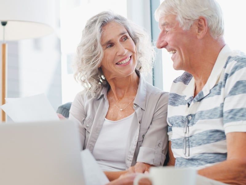 Older couple smiling, reviewing papers and using a laptop in a well-lit living room.