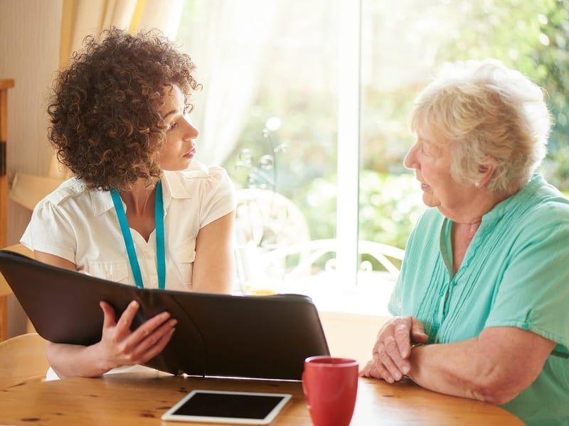 Woman in white shirt showing paperwork to an older woman in a turquoise shirt at a table near a window.