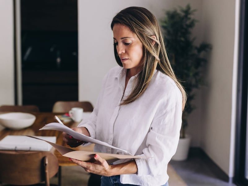 Woman in white shirt, looking at papers in a room with a wooden table and plants.