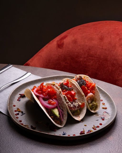 Tre tacos gourmet su un piatto grigio con guarnizione e salsa rosse, posizionati accanto a una sedia rossa.