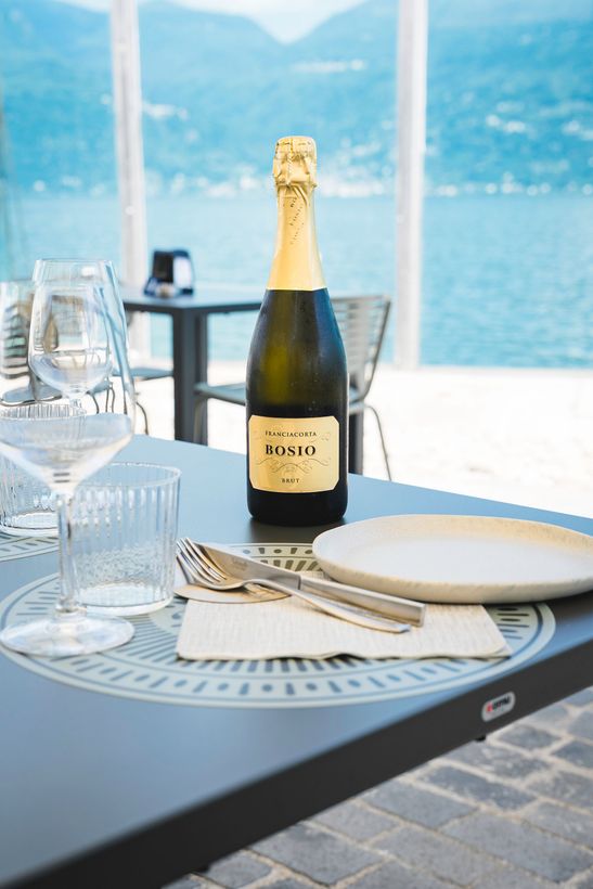 Bottiglia di champagne e tavola apparecchiata in riva a un lago con le montagne sullo sfondo.