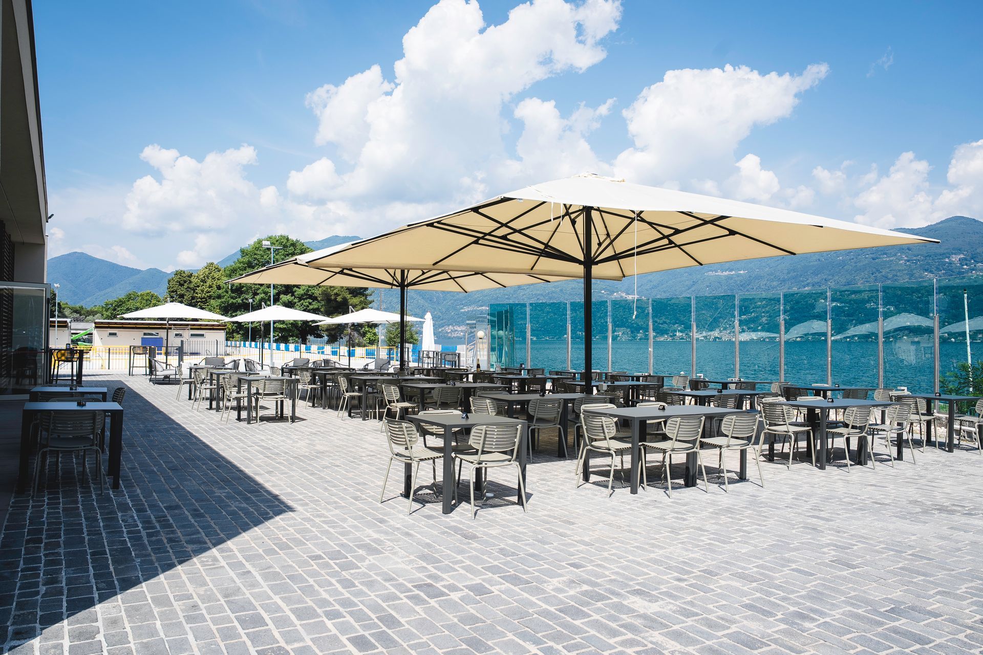 Patio esterno con tavoli e ombrelloni in riva a un lago, con le montagne sullo sfondo.