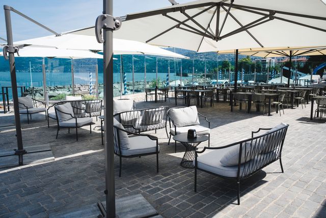 Terrazza esterna del caffè con sedie e tavoli neri e grandi ombrelloni bianchi con vista sull'acqua.