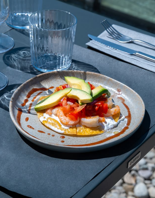 Toast con avocado e pomodoro su un piatto grigio, appoggiato su un tavolo all'aperto, con bicchieri e posate nelle vicinanze.