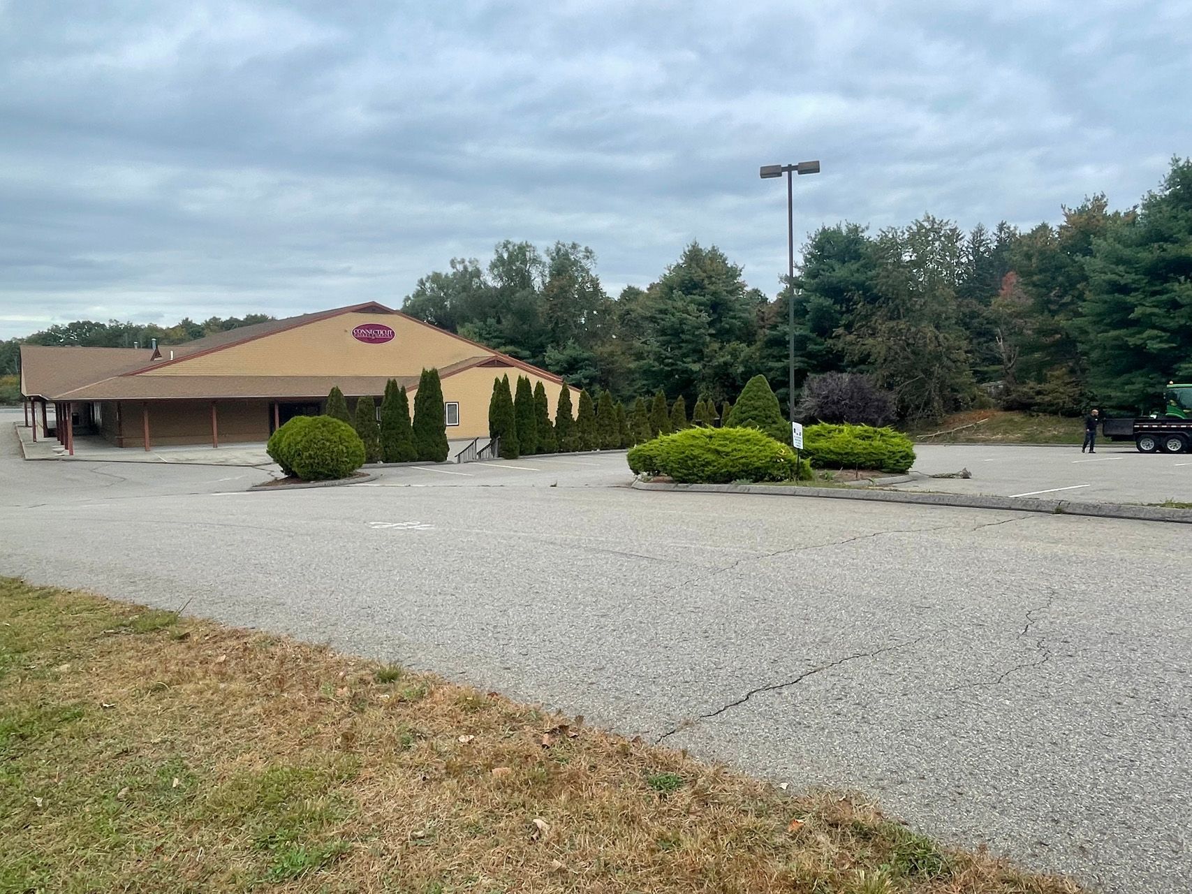 Empty parking lot beside a tan brick building with a red roof, bushes, and trees under an overcast sky