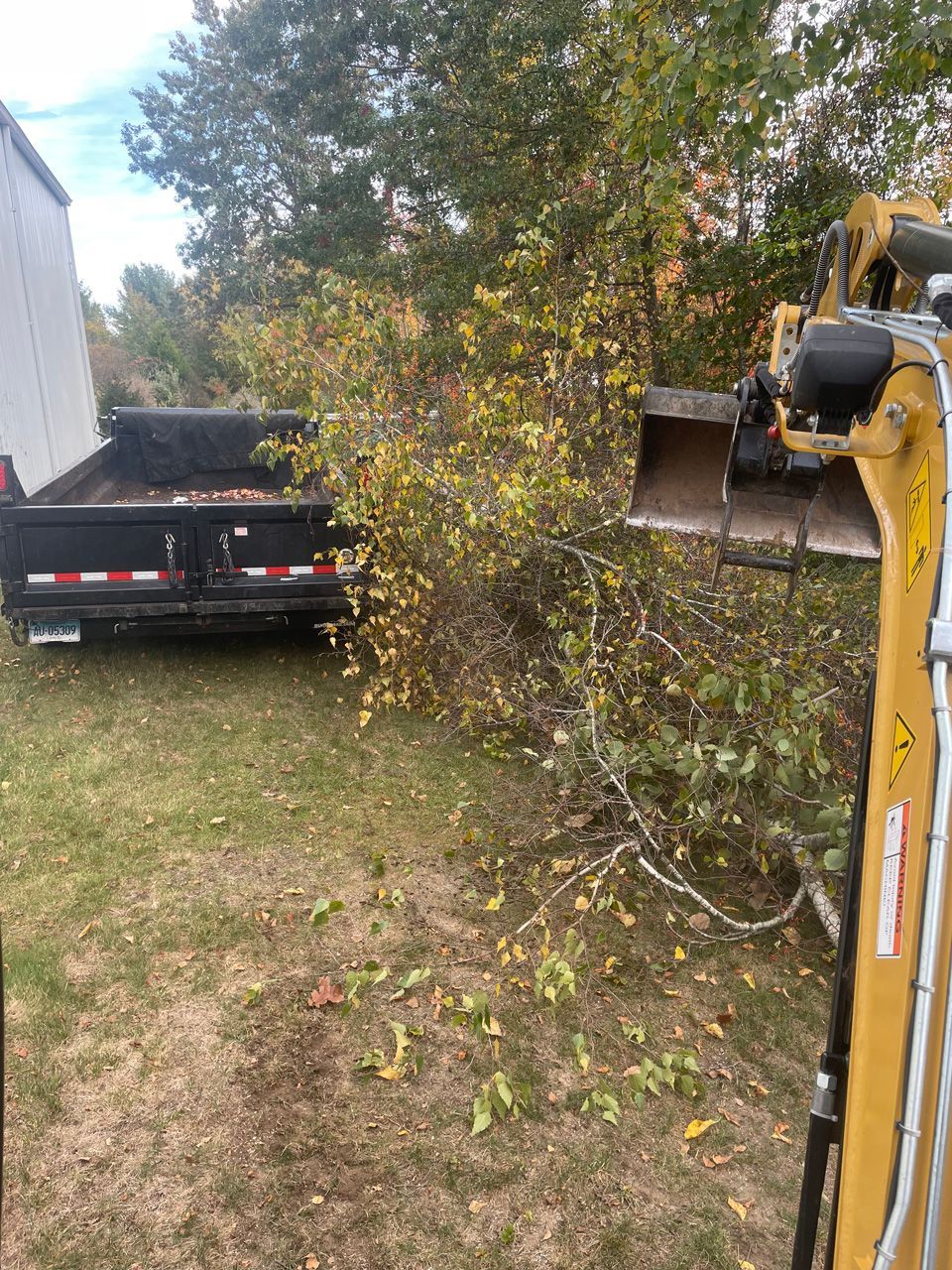 Yellow excavator clearing brush beside a black flatbed truck near a building.
