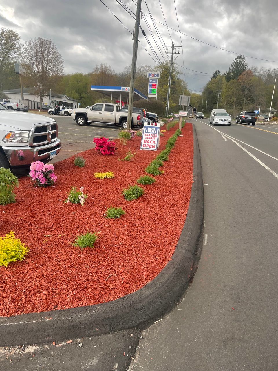 Red mulch flower bed with pink and yellow flowers beside a road and parking lot on a cloudy day