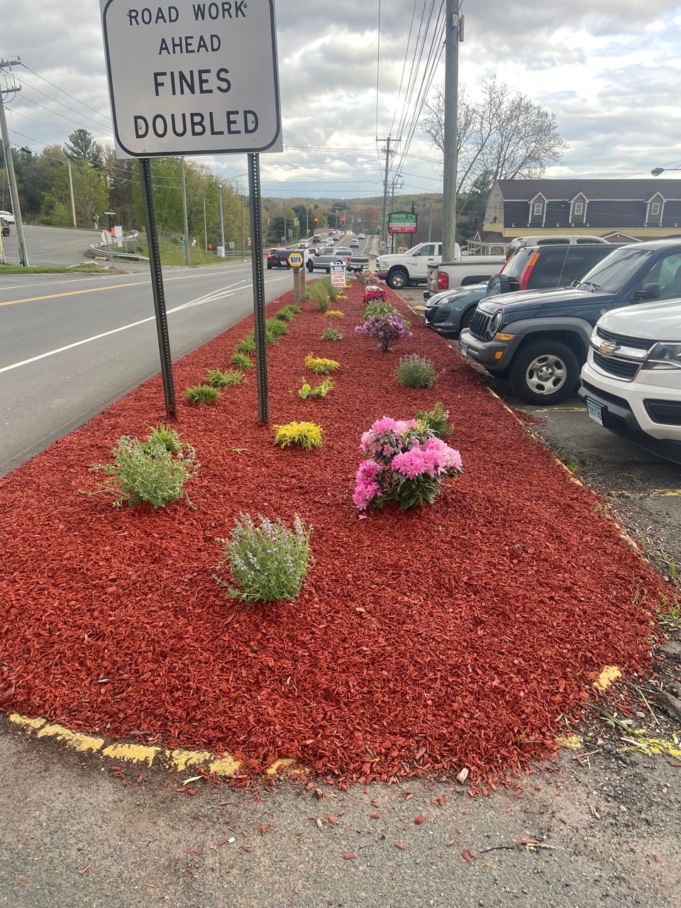 Mulched parking lot flower bed with pink blooms and a sign reading “Quads work ahead fines doubled.”
