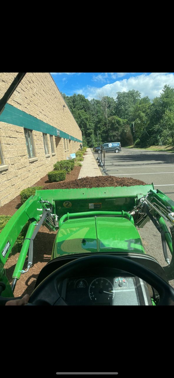Green tractor loader driving beside a tan brick building on a sunny street with trees ahead