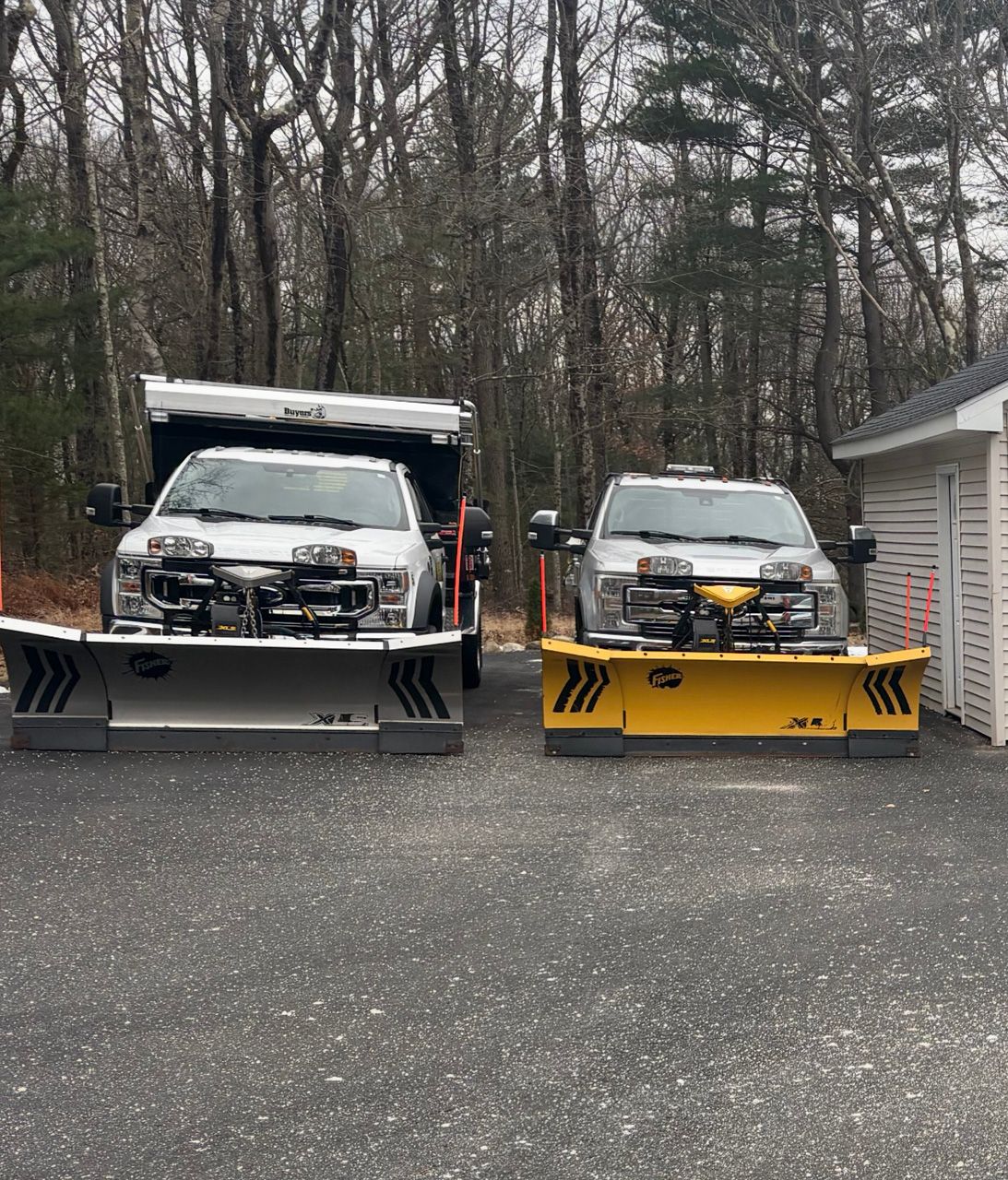Two snow plows parked in a driveway, one gray and one yellow, with a wooded house in the background.
