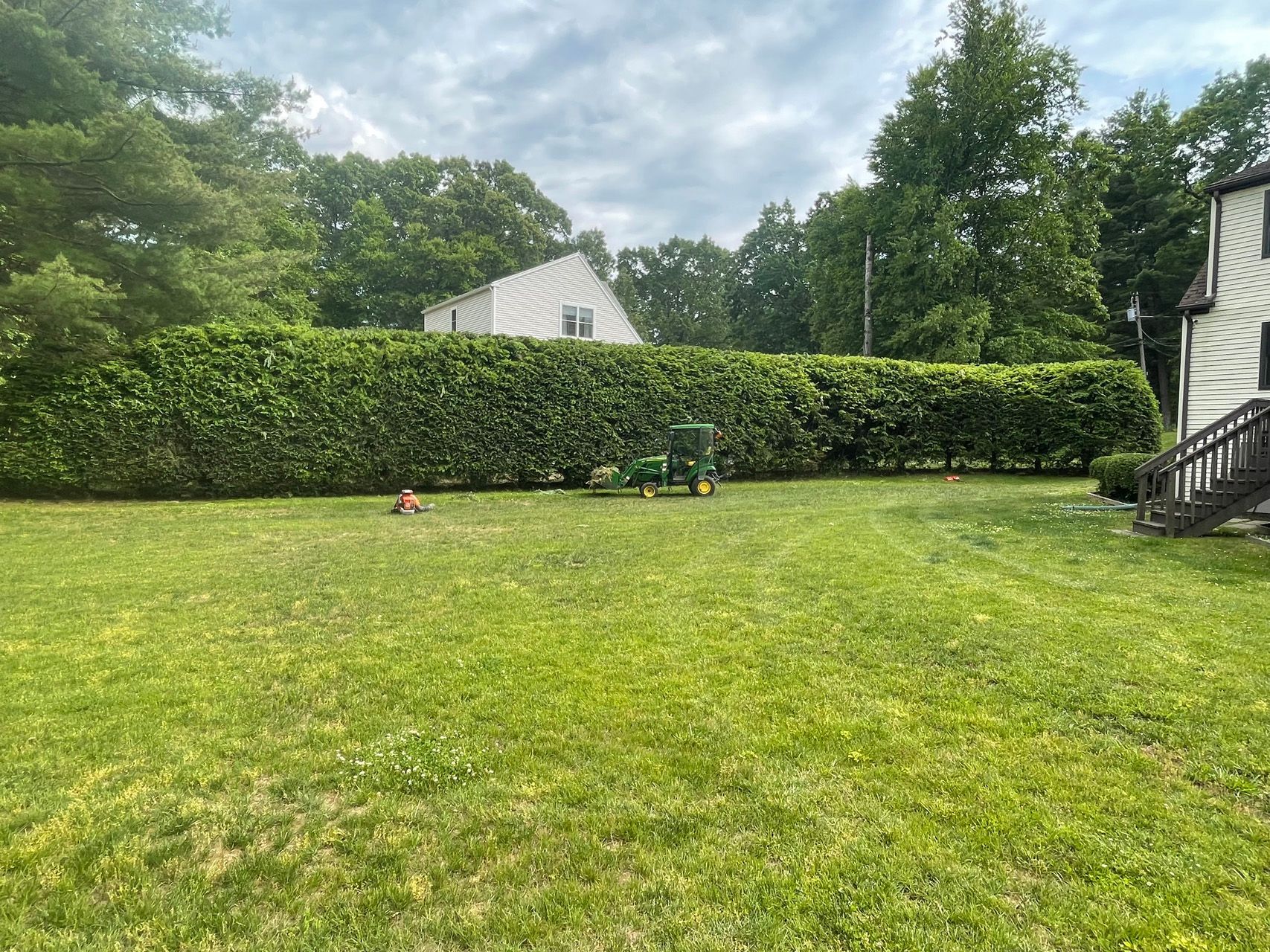 Grassy backyard with tall hedge, cloudy sky, and white house with deck on the right