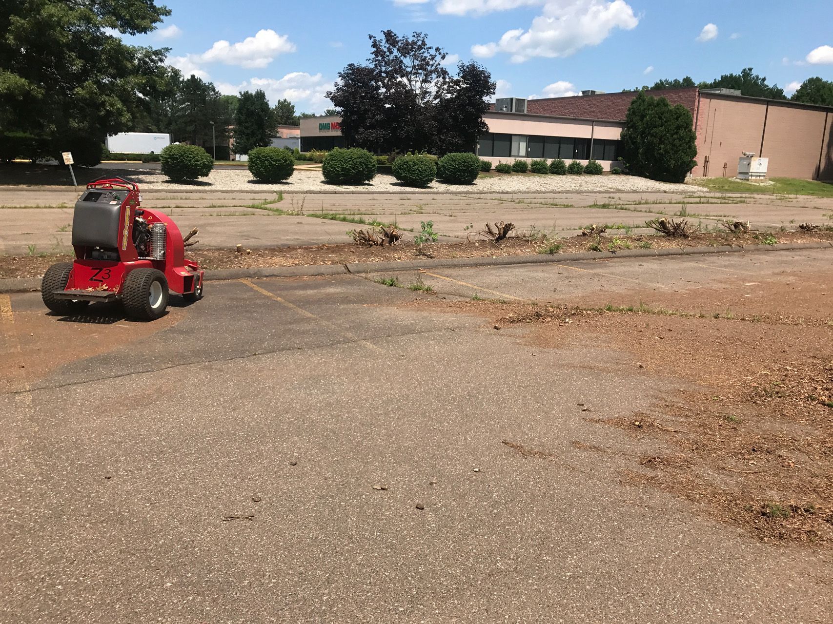 Small red ride-on mower on a gravel lot beside a building and trees under a partly cloudy sky