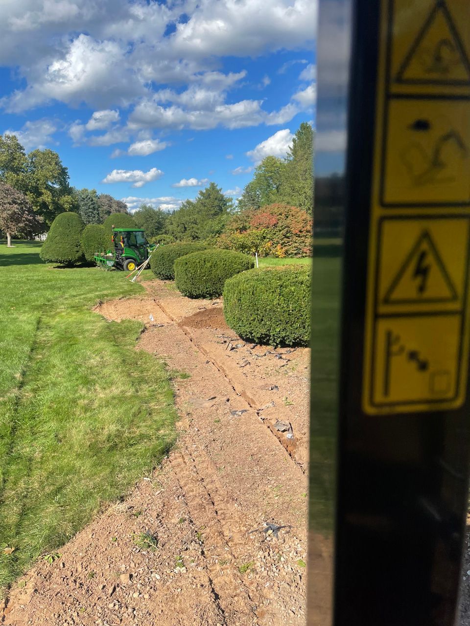 Sunny park path with trimmed bushes and blue sky, viewed beside a yellow warning sign.