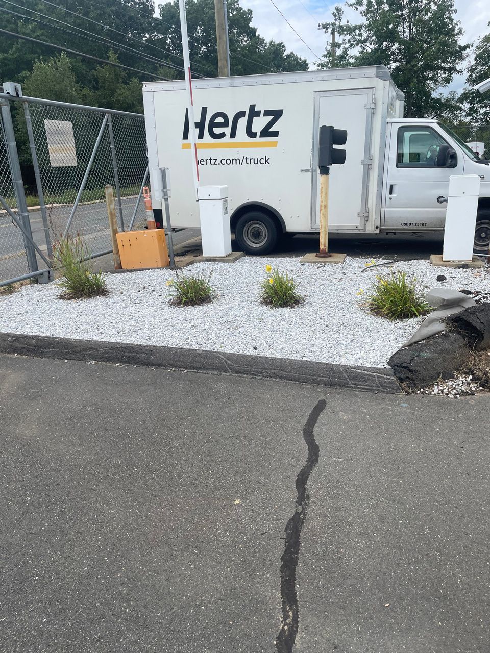 White Hertz box truck parked on a gravel lot beside a fence and utility pole.