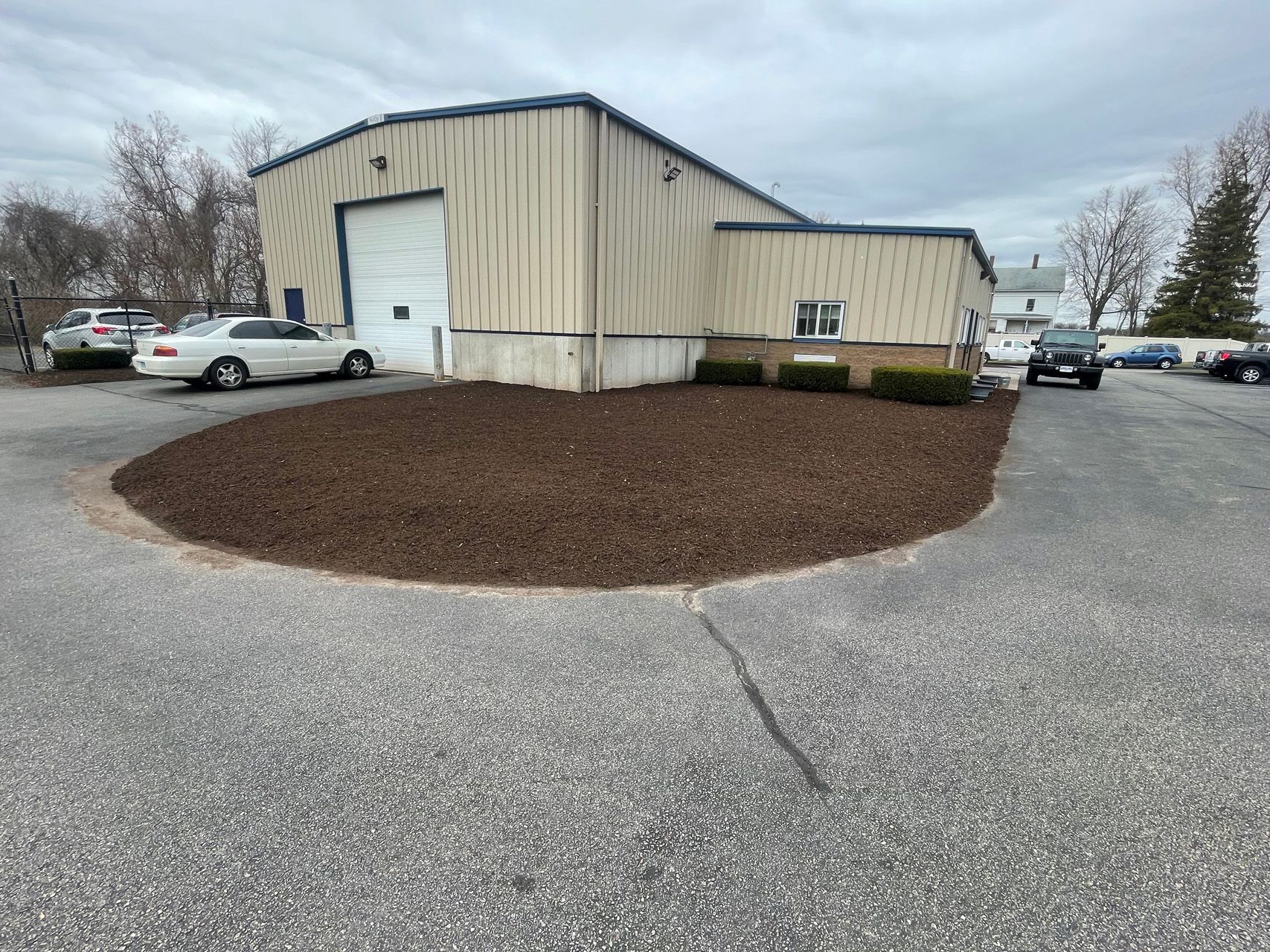 Industrial building with beige siding, parked cars, and a landscaped gravel lot under cloudy skies
