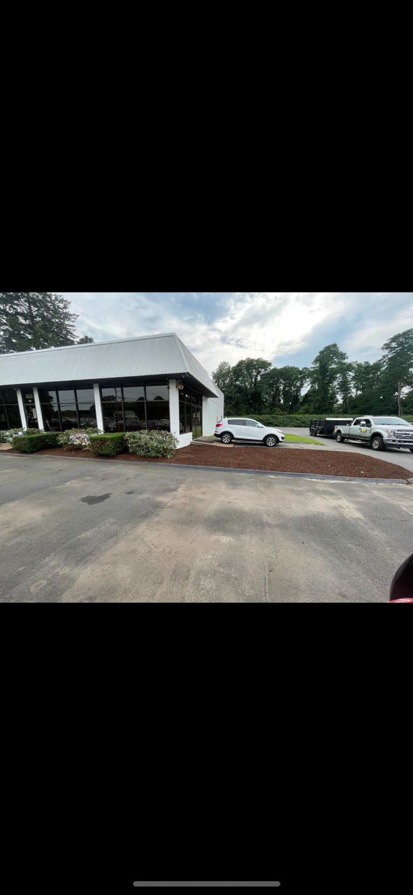 Small white building with a covered porch, parked car, and red gravel lot under a cloudy sky