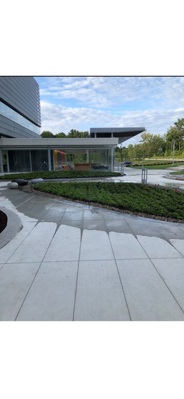 Modern building entrance with curved stone walkway, landscaped greenery, and blue sky.