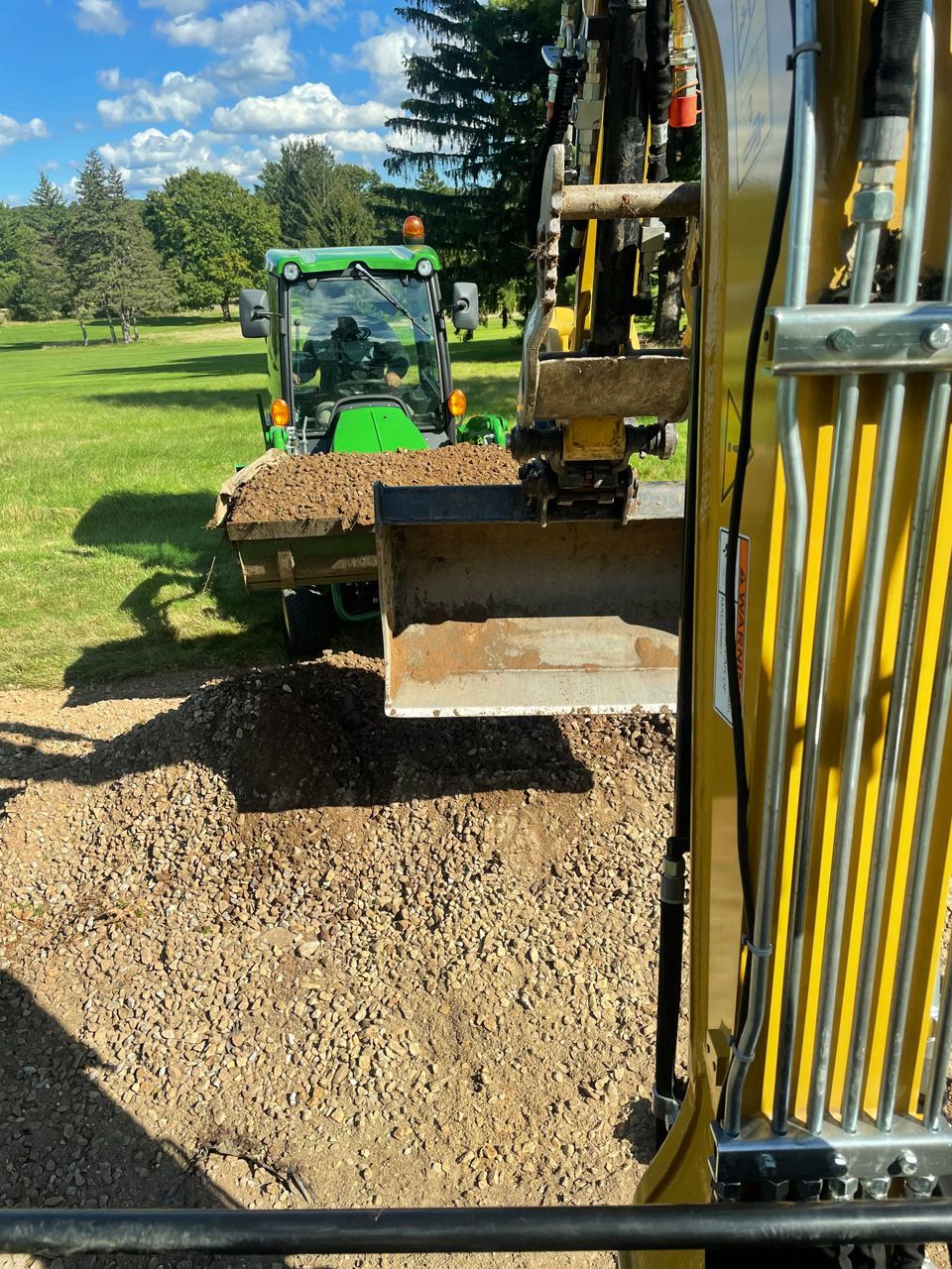 Excavator dumping gravel into a green dump trailer on a grassy work site under a blue sky