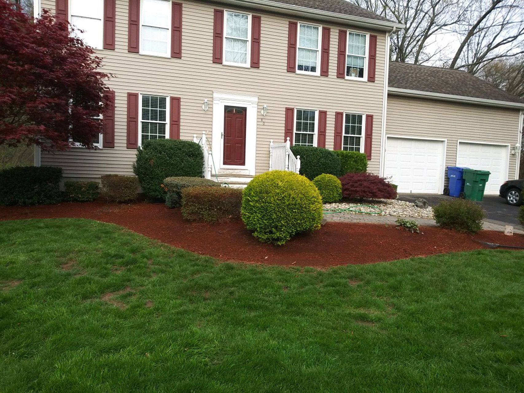 Suburban house with beige siding, red shutters, white garage, and landscaped front yard with shrubs and grass