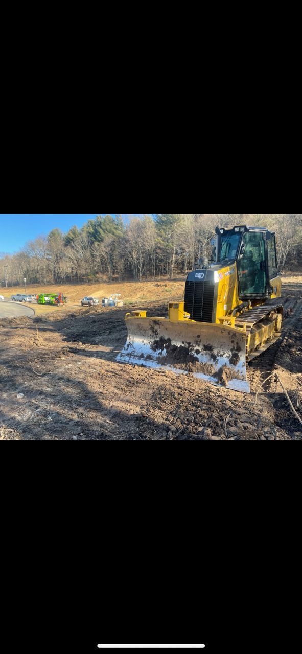 Yellow excavator working in a muddy field beside a water puddle, with trees in the background