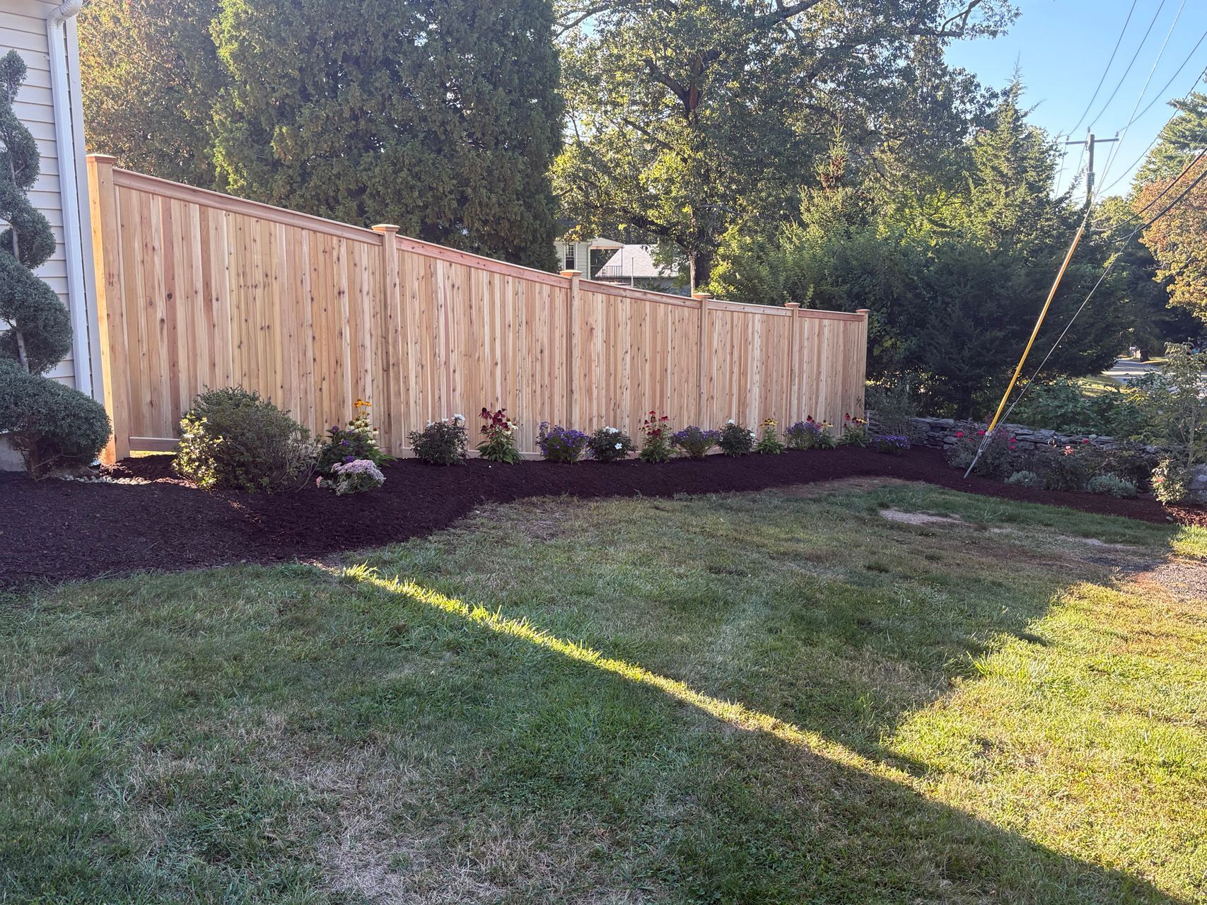 Wooden privacy fence with shrubs along a sloped backyard lawn in bright sunlight