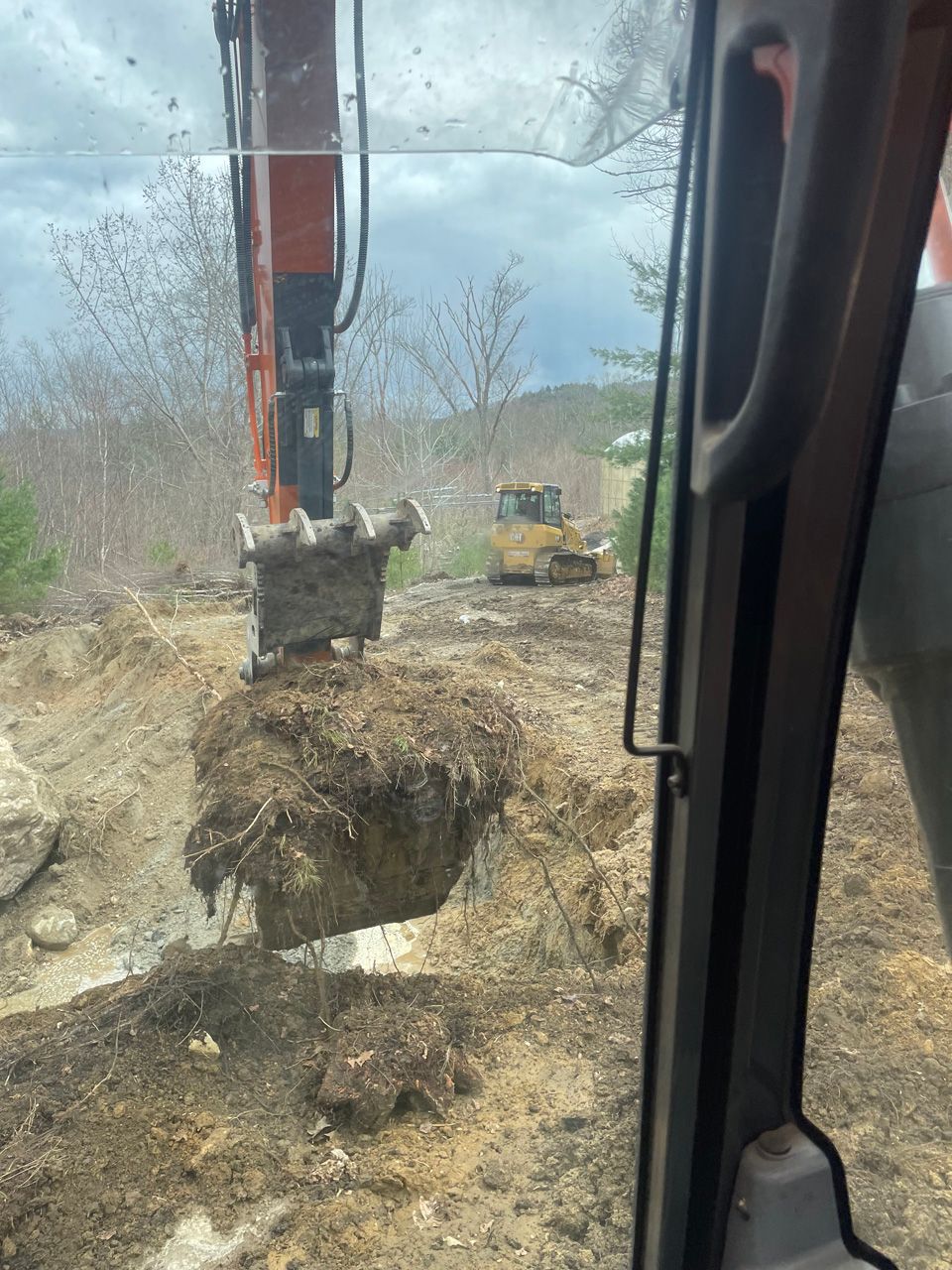 Excavator lifting a large dirt-root ball at a muddy construction site, viewed from inside another vehicle.