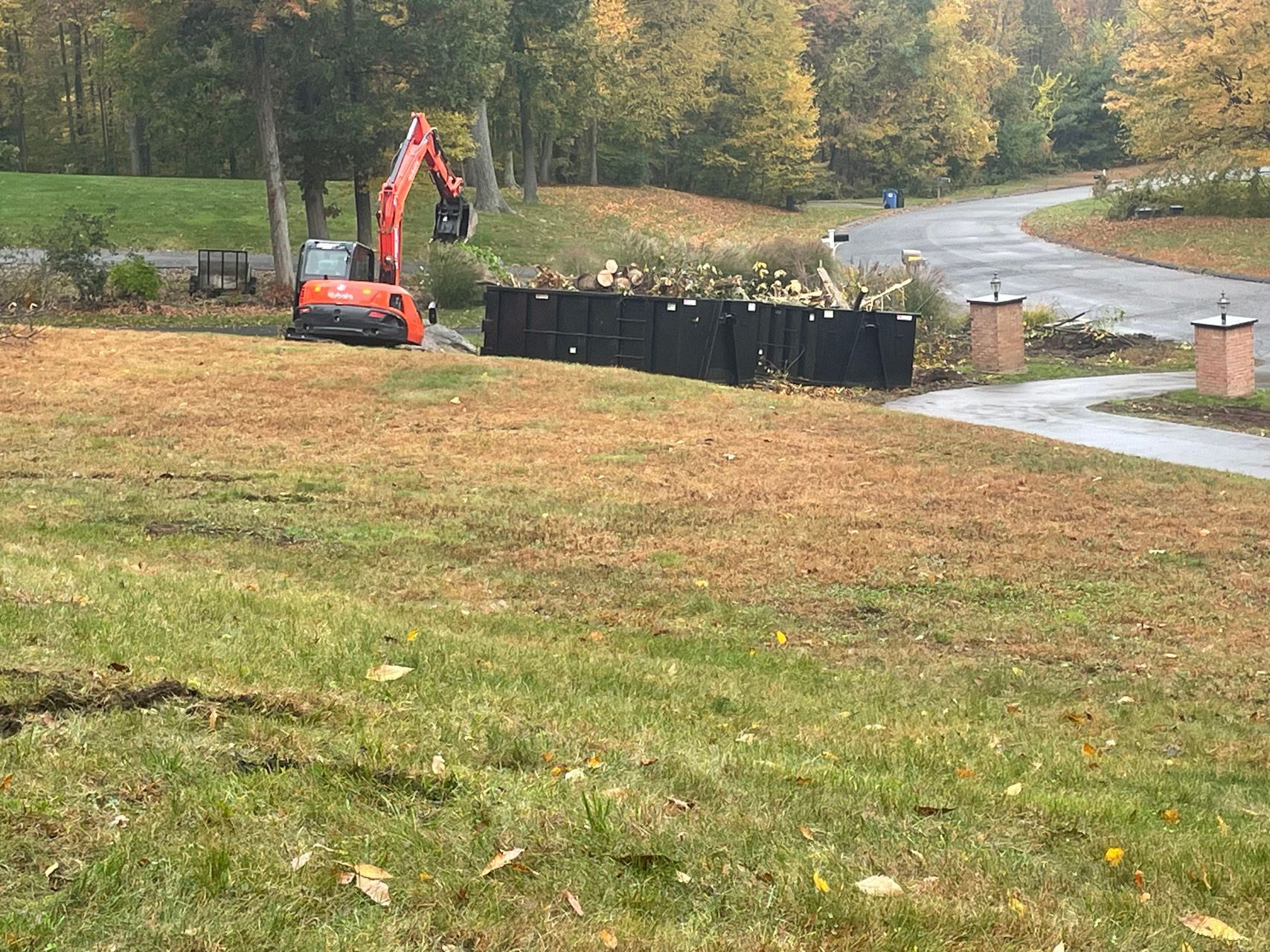Small orange excavator beside black silt fence on a grassy roadside slope in autumn.