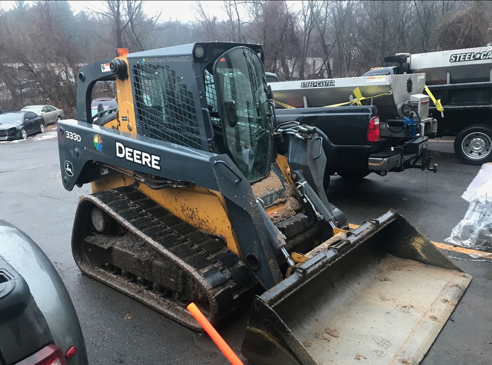 Yellow John Deere skid-steer loader with bucket in a snowy parking lot, towing equipment nearby